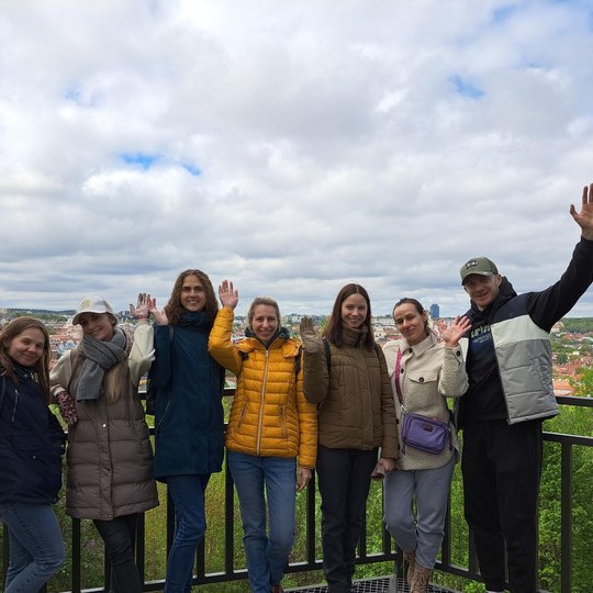 Grupė septynių besišypsančių žmonių pozuoja ant apžvalgos aikštelės su vaizdu į pavasarišką Vilniaus panoramą. Visi mojuoja rankomis, vilki šiltus drabužius, virš jų – debesuotas dangus su matomomis mėlyno dangaus properšomis.
