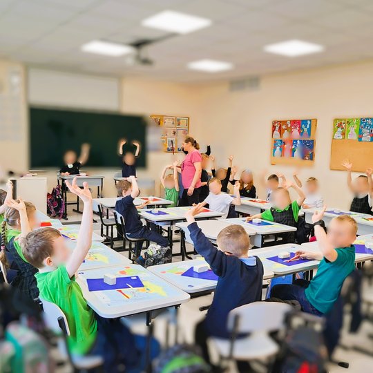 Pradinės mokyklos klasės nuotrauka, kurioje mokiniai sėdi prie savo suolų ir laiko rankose pakeltus molinius dirbinius. Vaikai vilki mokyklines uniformas, klasėje šviesu, pro langus sklinda dienos šviesa. Ant suolų padėtos spalvotos lentelės darbui, o fon