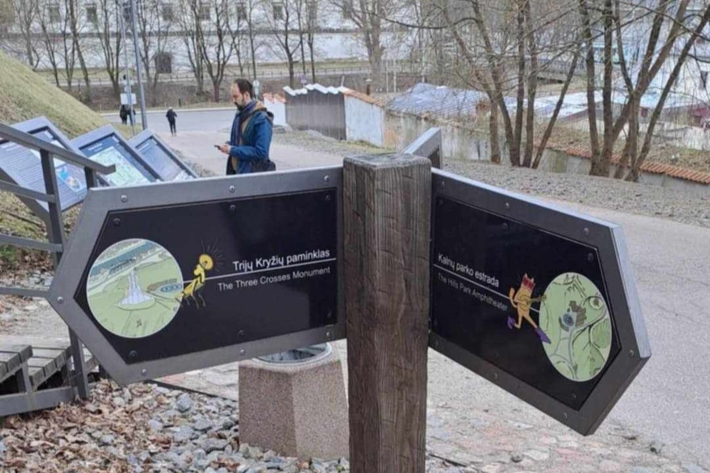 Rodyklės formos informaciniai stendai Vilniaus Kalnų parke rodo kryptis link Trijų Kryžių paminklo (kairėje) ir Kalnų parko estrados (dešinėje); fone matyti praeiviai, informaciniai stendai, tvora, medžiai be lapų.
