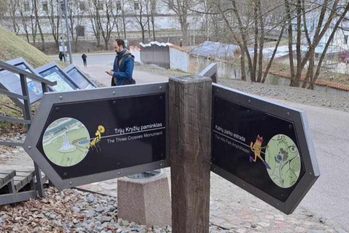 Rodyklės formos informaciniai stendai Vilniaus Kalnų parke rodo kryptis link Trijų Kryžių paminklo (kairėje) ir Kalnų parko estrados (dešinėje); fone matyti praeiviai, informaciniai stendai, tvora, medžiai be lapų.