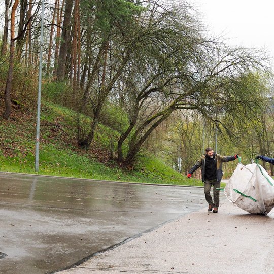 Du žmonės tempia didelį baltą šiukšlių maišą šlapiu asfaltuotu keliu, apsuptu pavasarinių medžių ir žolės. Abu dėvi lietui tinkamus drabužius ir pirštines, o veiduose matyti gera nuotaika. Fone – miškingas šlaitas ir žaliuojanti gamta.