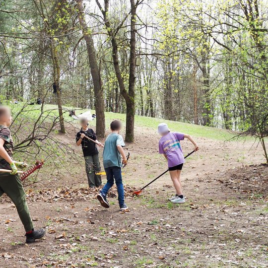 Miške grupė vaikų aktyviai dalyvauja aplinkos tvarkymo veikloje – grėbia lapus, nešasi darbo įrankius. Aplink juos – pavasariškai sužaliavę medžiai, ant žemės – pernykščiai lapai. Vaikai dėvi pirštines, naudoja grėblius.