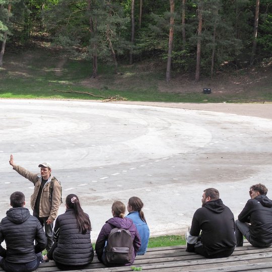 Vyras su kuprine stovi priešais sėdinčią grupę žmonių ir gestikuliuodamas aiškina kažką lauko amfiteatro aikštelėje. Fone matyti miško pakraštys ir betoninis apskritimų raštais išmargintas grindinys.