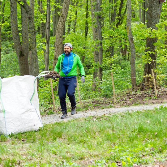 Miško take einantis vyras su žalia striuke neša glėbį sausų lapų ir ruošiasi juos mesti į baltą maišą. Aplinkui veši žaluma, o fone stovi seni medžiai ir apsauginė tvorelė.