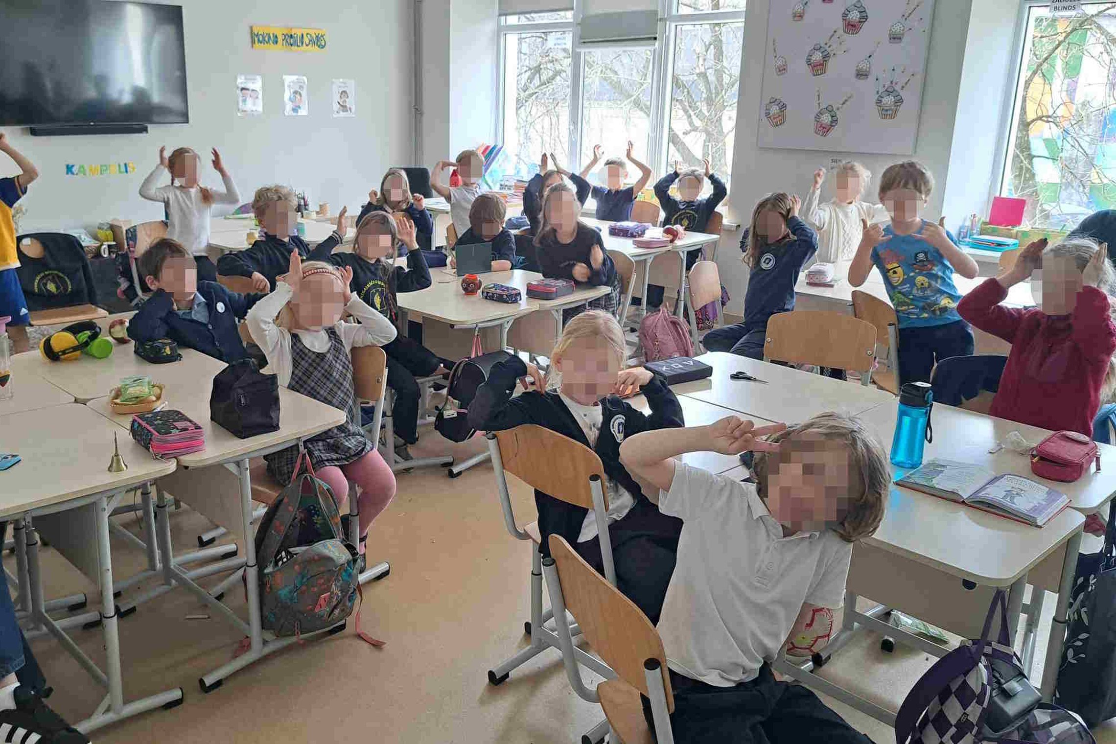 Klasės mokiniai aktyviai dalyvauja klasės veikloje, keldami rankas aukštyn. Kambarys šviesus, stalai sustatyti eilėmis, ant kurių matomi mokykliniai reikmenys ir kuprinės. Fone matosi švieslentė ir mokyklinės dekoracijos.