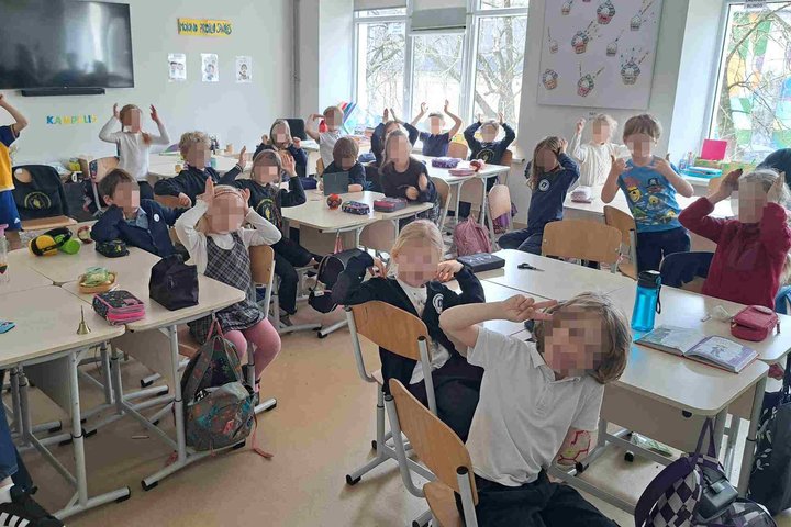 Klasės mokiniai aktyviai dalyvauja klasės veikloje, keldami rankas aukštyn. Kambarys šviesus, stalai sustatyti eilėmis, ant kurių matomi mokykliniai reikmenys ir kuprinės. Fone matosi švieslentė ir mokyklinės dekoracijos.