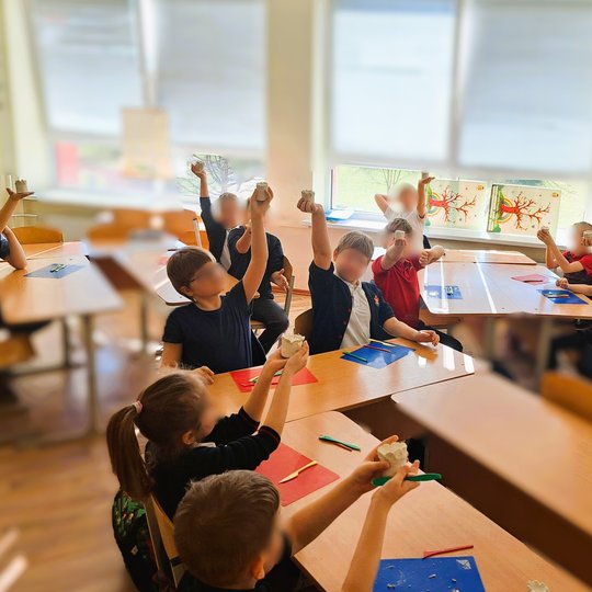 Vaikai klasėje sėdi prie stalų ir iškėlę rankose rodo savo nulipdytas pilaites iš molio. Ant langų kabo rudeniniai piešiniai, o ant stalų – padėkliukai ir įrankiai kūrybinei veiklai.