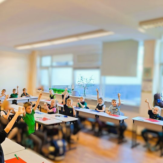 Pradinės mokyklos klasės nuotrauka, kurioje mokiniai sėdi prie savo suolų ir laiko rankose pakeltus molinius dirbinius. Vaikai vilki mokyklines uniformas, klasėje šviesu, pro langus sklinda dienos šviesa. Ant suolų padėtos spalvotos lentelės darbui, o fon