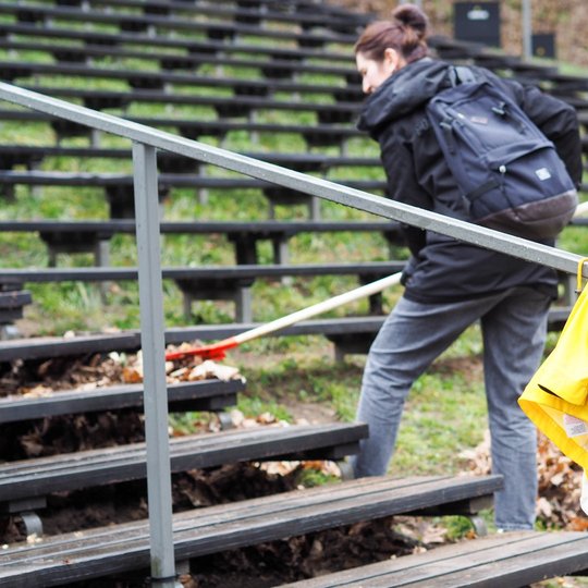 Lauko estradoje moteris su juodu kupriniu grėbia lapus nuo suolų, naudodama oranžinį grėblį. Ji stovi pasilenkusi tarp medinių suolų, vilkėdama tamsią striukę ir pilkas kelnes. Priekyje, ant metalinio turėklo, pakabinta ryškiai geltona lietaus striukė.