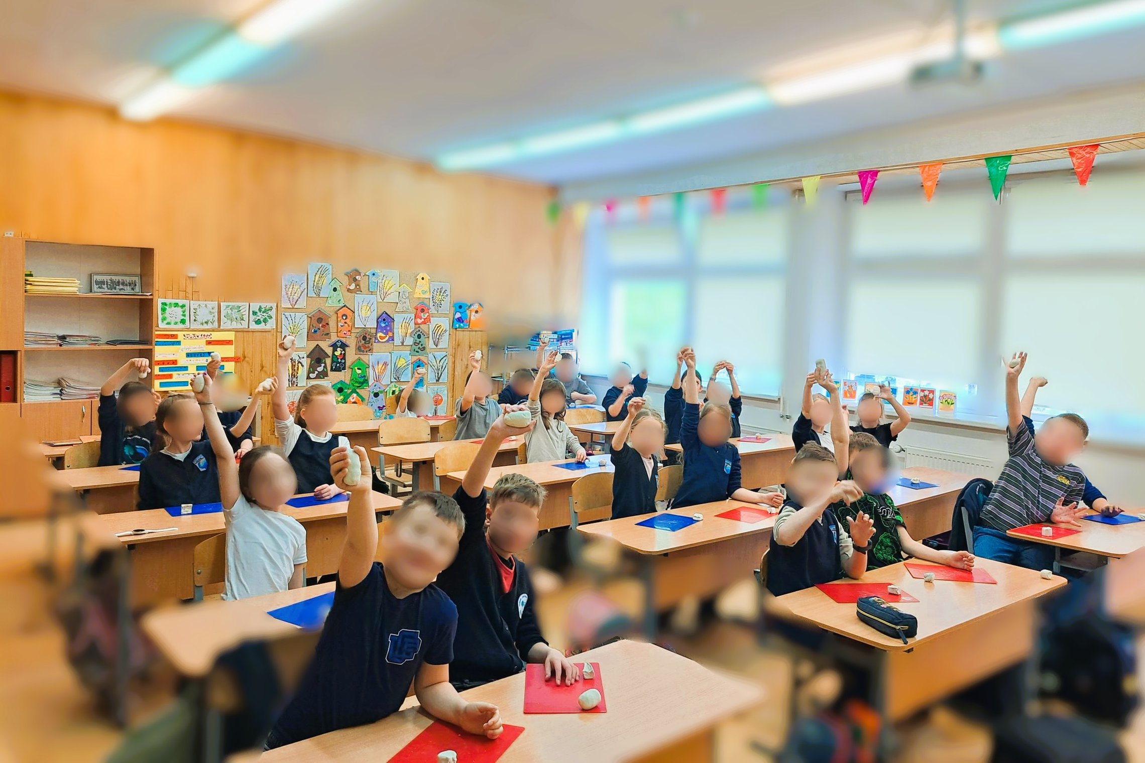 Klasė pilna vaikų. Rankose jie laiko figūrėles. Aplinka šviesi, klasė pilna mokymosi priemonių