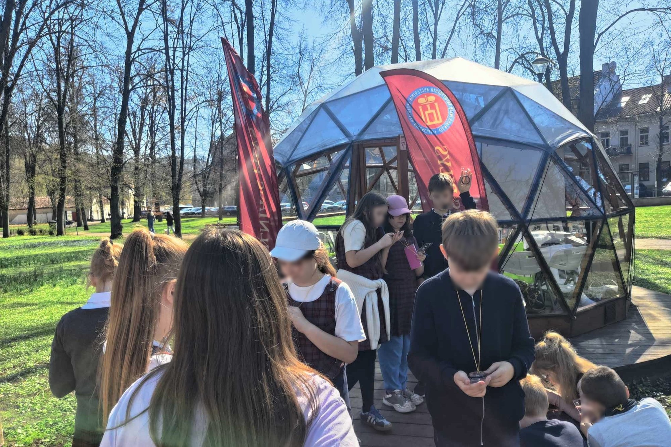 Trečiokai su kompasais tyrinėja Vilniaus pilių teritoriją prie kupolo. Aplink matyti pavasariniai medžiai, žolė ir senamiesčio pastatai. Mokiniai susikaupę stebi kompasų rodykles ir užrašo pastebėjimus.