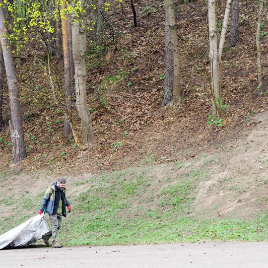 Vyras traukia baltą šiukšlių maišą asfaltuotu taku šalia miškingo šlaito. Aplink auga pavasarėjantys medžiai, žolė ir dar likę pernykščiai lapai.