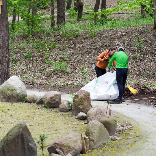 Du žmonės šalia miško tako pila surinktus lapus į baltą maišą, stovintį ant žemės. Priekyje matomas nedidelis akmenų ratas, o aplinkui – miško šlaitas ir žaliuojantys medžiai.