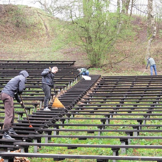 Keli žmonės valo lauko estrados suolus – grėbia lapus ir renka juos į krūvas tarp eilių. Aplink matyti žaliuojanti žolė, medžiai su pavasariniais pumpurais ir šlaitas fone.
