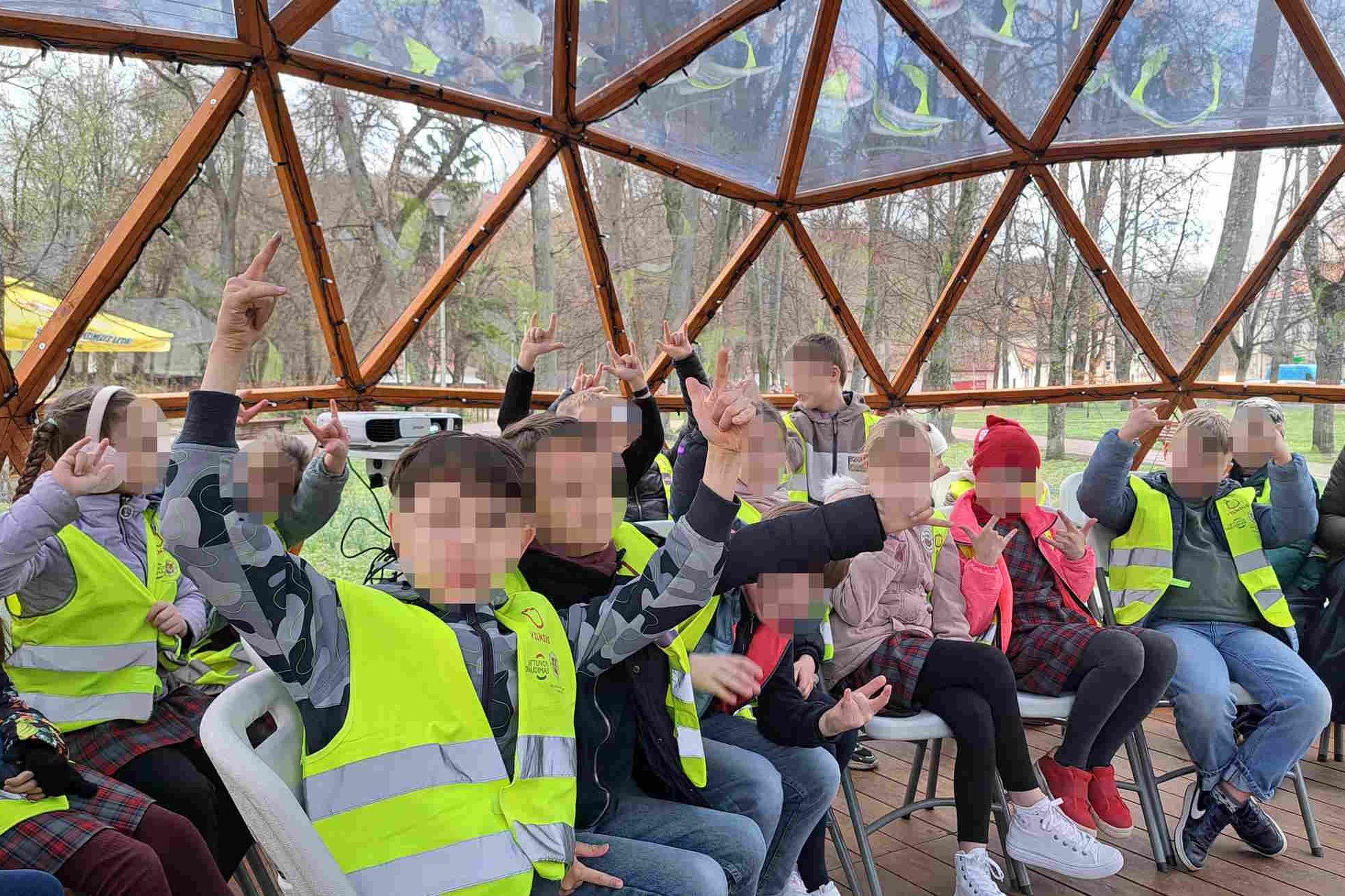 Grupė vaikų su ryškiai geltonomis liemenėmis sėdi kupolo formos medinėje patalpoje, keli iš jų pakėlę rankas rodo roko simbolį. Fone matyti pavasariški medžiai ir miesto parkas. Edukacija vyksta viduje po skaidriu kupolu.