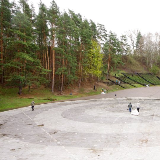 Didelėje lauko estradoje, apsuptoje miškingų kalvų ir medinių žiūrovų suoliukų, žmonės atlieka tvarkymo darbus – renka šiukšles, neša maišus. Priekyje matyti keli asmenys su šiukšlių maišais, tolėliau – daugiau talkininkų, pasklidusių po visą teritoriją.