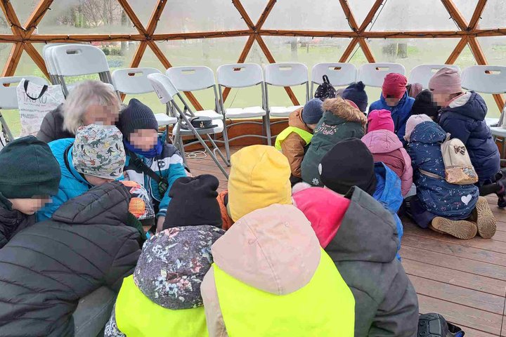 Nuotraukoje matoma grupė mažų vaikų, susibūrusių ratu ant medinių grindų šiltnamio formos kupole. Jie vilki šiltus lauko drabužius ir kepures, kai kurie nešioja ryškiaspalvius liemenes. Aplinkui stovi tuščos baltos kėdės. Vaikai įsitraukę į bendrą veiklą,