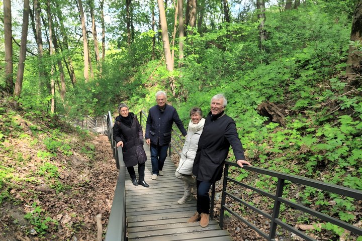 Keturi žmonės eina mediniais laiptais kalno šlaite. Jie šypsosi, sustoję trumpam pasifotografuoti tarp pavasarį sužaliavusių medžių ir tankios žalumos.