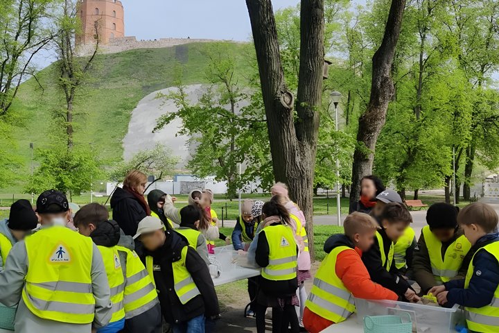 Vaikai su ryškiais geltonomis liemenėmis dalyvauja edukacinėje veikloje lauke, prie stalų su priemonėmis. Veiksmas vyksta prie Gedimino kalno papėdės, fone matoma Gedimino pilies bokšto viršūnė ir žalias šlaitas.