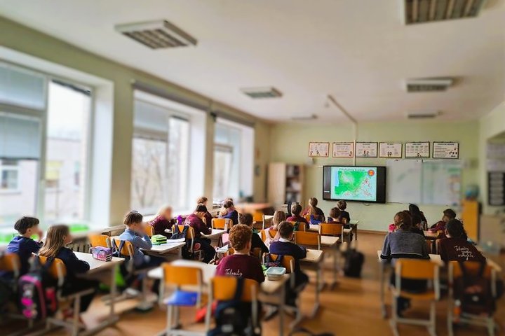 Pradinukai sėdi klasėje prie savo suolų ir žiūri į interaktyvią lentą, kurioje rodoma žemėlapio skaidrė. Klasė šviesi, su dideliais langais ir šviesiomis sienomis, ant kurių kabo įvairūs edukaciniai plakatai. Vaikai dėvi uniforminius marškinėlius.
