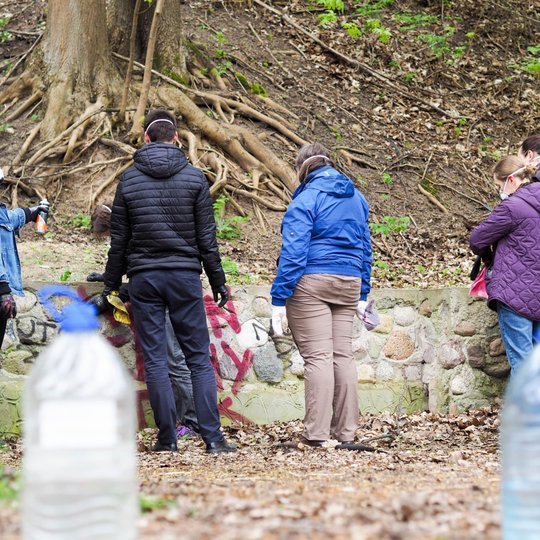 Grupė žmonių stovi prie akmeninės sienelės, išteptos grafičiais, ir ruošiasi ją valyti. Kai kurie laiko purškiklius ir šluostes, visi dėvi apsaugines kaukes.