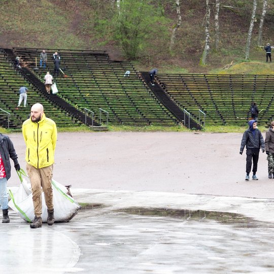 Prie lauko estrados du vyrai tempia didelį šiukšlių maišą, o už jų, tarp suolų, matyti daugiau žmonių, užsiimančių tvarkymo darbais. Aplinka apsupta medžių, o žemė ir suolai atrodo šlapi nuo lietaus.