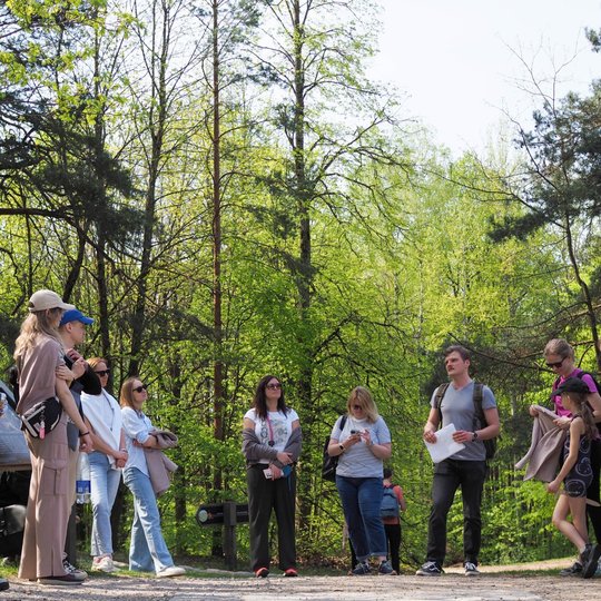 Miško apsuptyje stovi žmonių grupė, išklausanti kalbančio vyro, kuris rankoje laiko lapus. Visi apsirengę laisvalaikio drabužiais, o aplink žaliuoja pavasariniai medžiai.