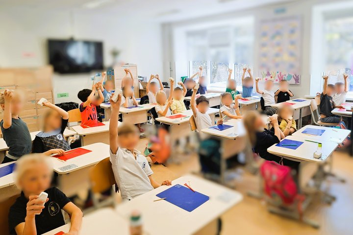 Nuotraukoje matome pradinių klasių mokinius klasėje. Vaikai sėdi prie stalų, pakėlę rankas aukštyn, laikydami kažką rankose – tai modelinas. Klasėje šviesu, pro didelius langus sklinda dienos šviesa. Ant sienų kabinami plakatai, vienas jų – „MŪSŲ GIMTADIE