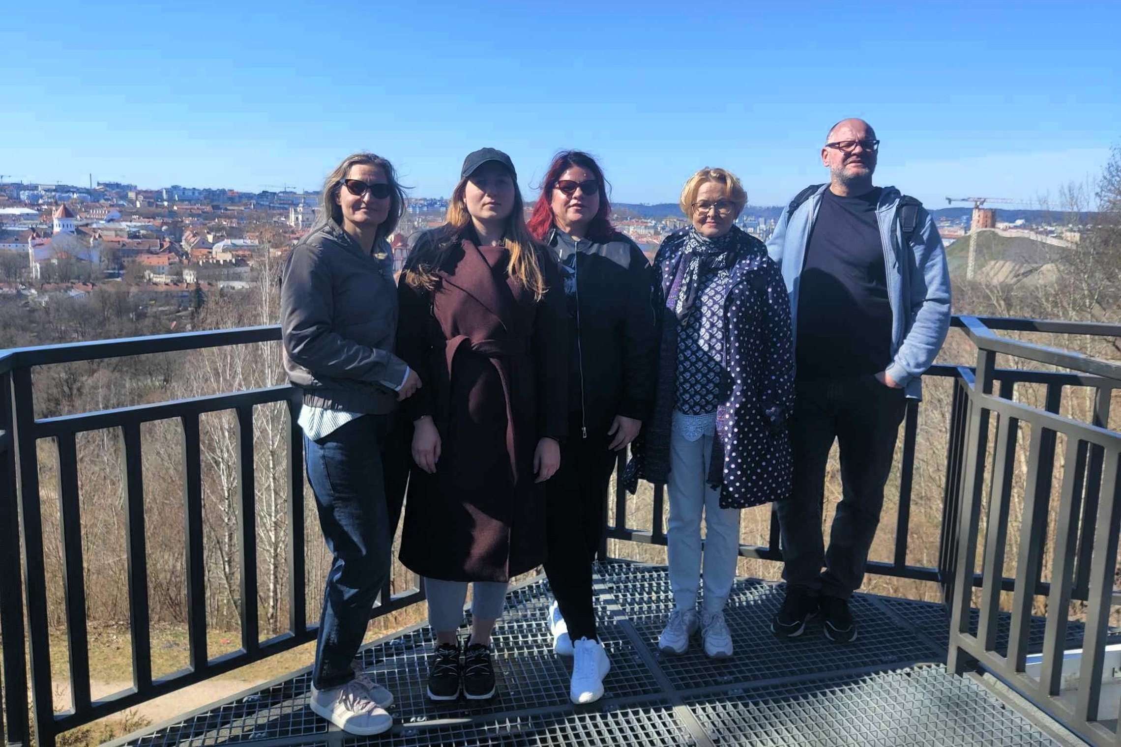 Nuotraukoje – penki žmonės stovi ant apžvalgos Altanos bokštelio aikštelės, nuo kurios atsiveria Vilniaus senamiesčio panorama. Fone matyti raudoni senamiesčio stogai, bažnyčių bokštai, o tolumoje – Gedimino pilies bokštas ant kalno. Diena giedra, dangus