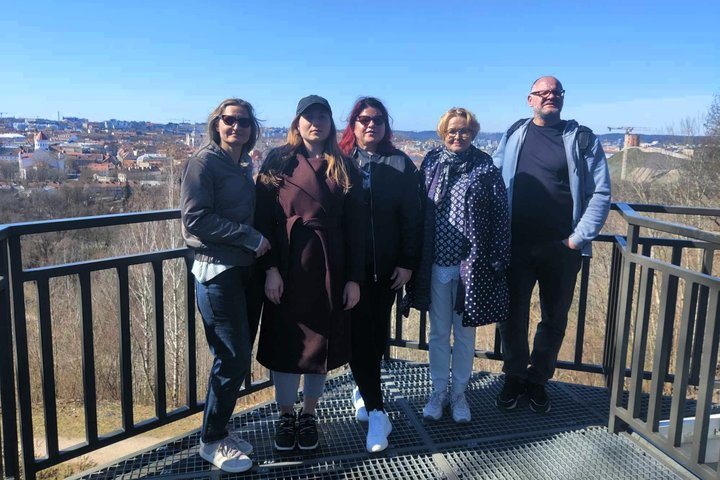 Nuotraukoje – penki žmonės stovi ant apžvalgos Altanos bokštelio aikštelės, nuo kurios atsiveria Vilniaus senamiesčio panorama. Fone matyti raudoni senamiesčio stogai, bažnyčių bokštai, o tolumoje – Gedimino pilies bokštas ant kalno. Diena giedra, dangus