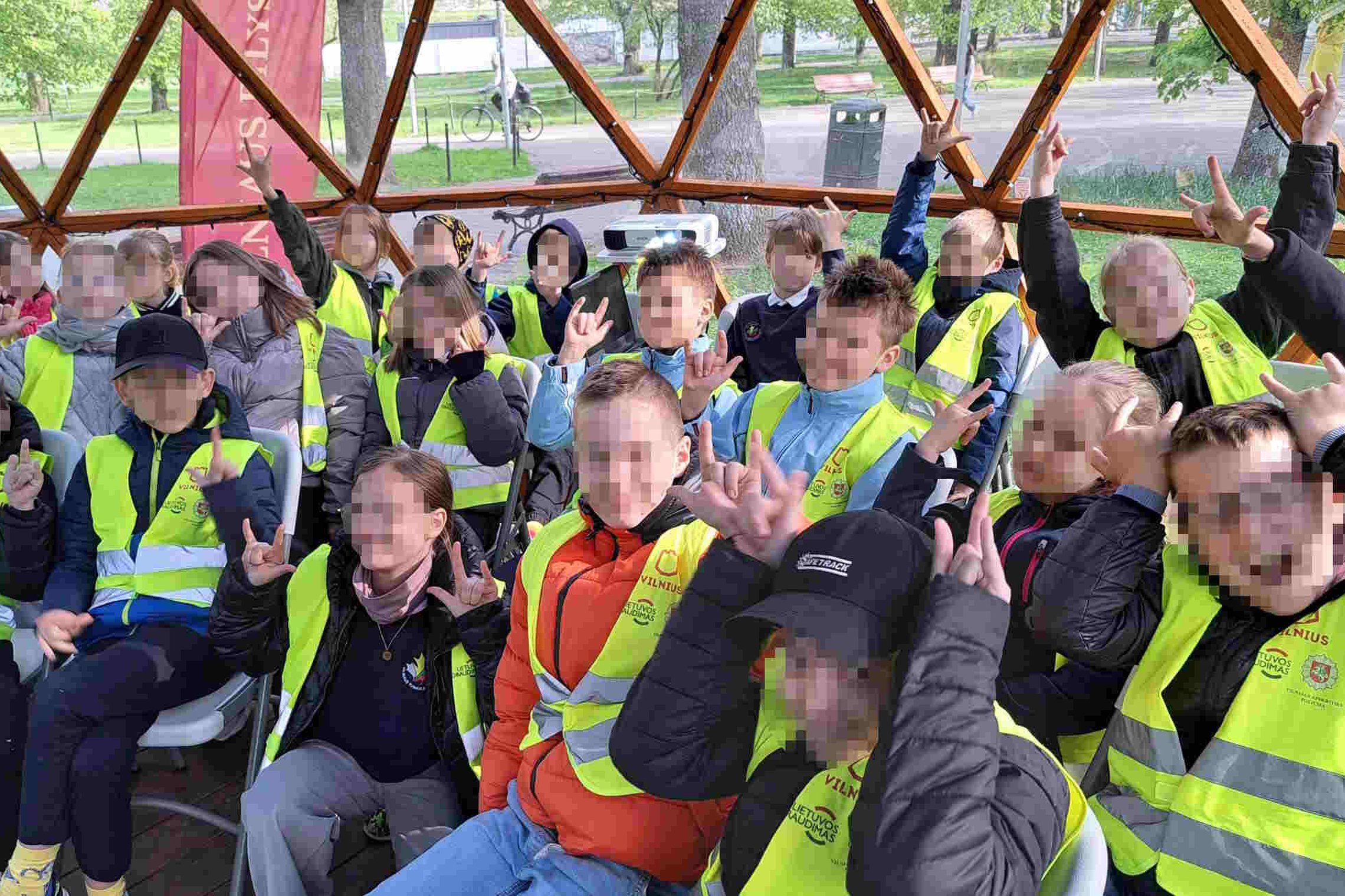Grupė trečiokų su ryškiais geltonais atšvaitiniais liemenėmis sėdi kupole ir rodo roko ženklus. Už jų pro langus matyti parkas ir pavasariška žaluma.