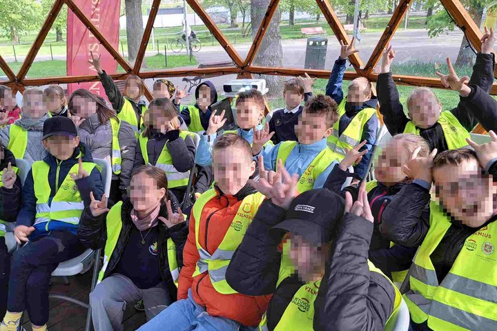 Grupė trečiokų su ryškiais geltonais atšvaitiniais liemenėmis sėdi kupole ir rodo roko ženklus. Už jų pro langus matyti parkas ir pavasariška žaluma.