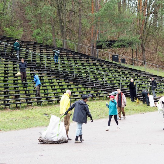 Didelė žmonių grupė dalyvauja talkoje prie lauko estrados, renka šiukšles ir grėbia lapus tarp medinių suolų. Priekyje keli žmonės neša ar tempia baltus šiukšlių maišus, o fone matyti miškingas šlaitas.
