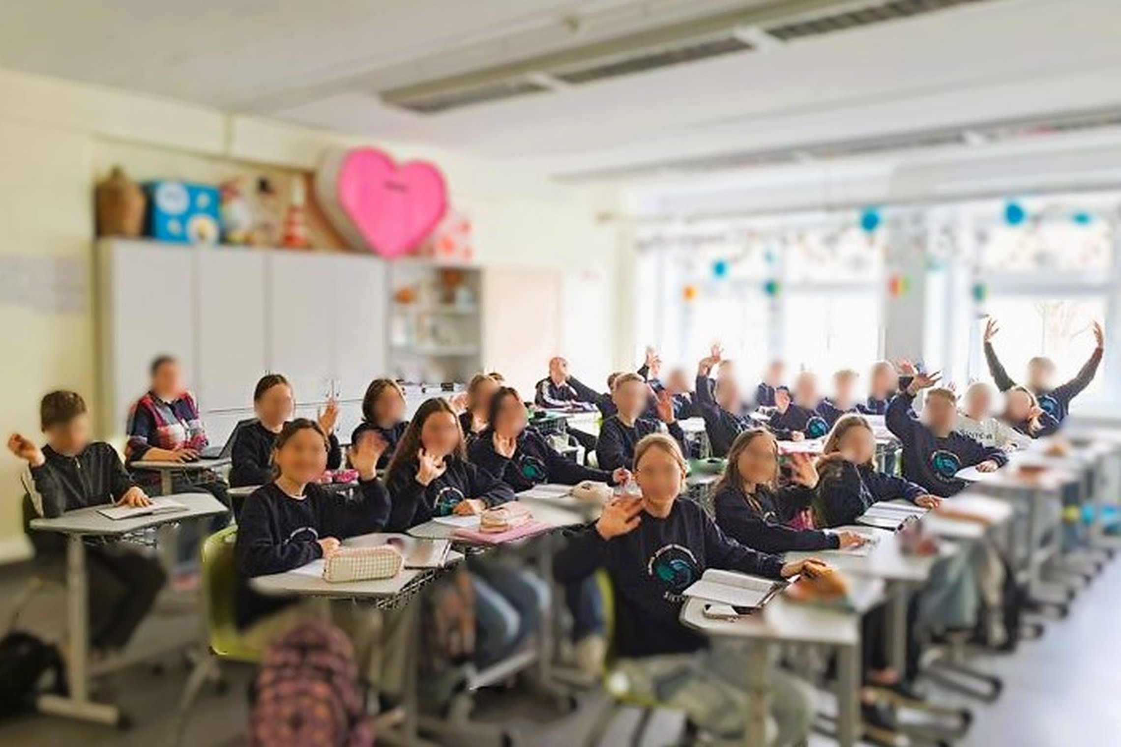 Nuotraukoje 27 žmonės, 26 vaikai ir 1 suaugęs. Tai klasės nuotrauka, kurioje mokiniai sėdi prie stalų, daugelis jų pakėlę rankas, mojuoja į objektyvą. Visi vilki vienodus džemperius su mokyklos logotipo emblema. Kambarys šviesus ir išpuoštas. Už mokinių, 