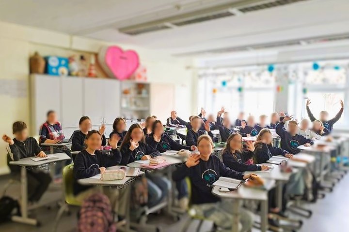 Nuotraukoje 27 žmonės, 26 vaikai ir 1 suaugęs. Tai klasės nuotrauka, kurioje mokiniai sėdi prie stalų, daugelis jų pakėlę rankas, mojuoja į objektyvą. Visi vilki vienodus džemperius su mokyklos logotipo emblema. Kambarys šviesus ir išpuoštas. Už mokinių,