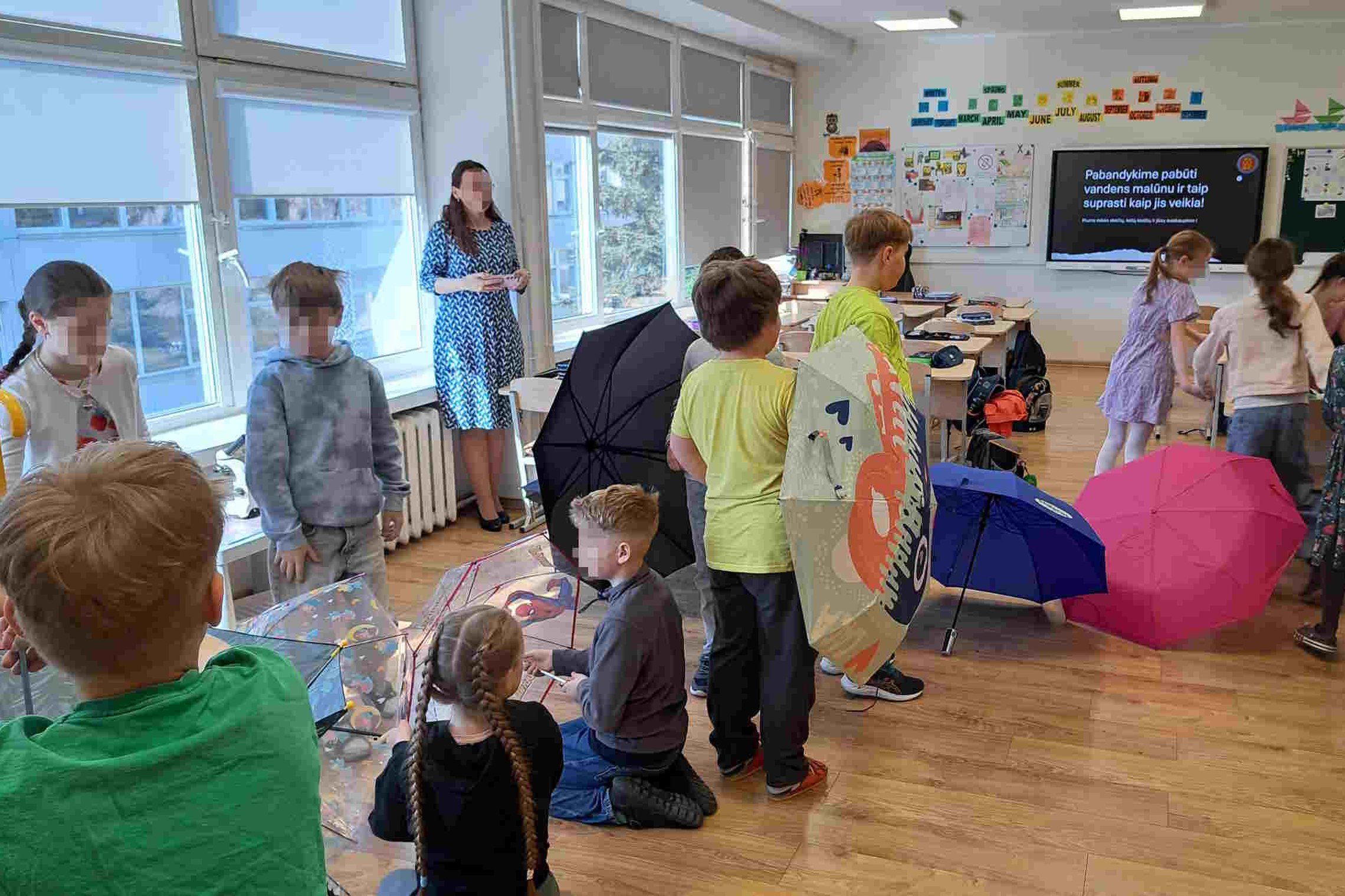 Nuotraukoje – pradinukai klasėje, dalyvaujantys pamokoje apie vandens malūną. Vaikai su skėčiais vaidina malūno mechanizmus. Prie lango stovi mokytoja, o lentoje parašyta: „Pabandykime pabūti vandens malūnu ir taip suprasti kaip jis veikia!“ Aplinka kūryb
