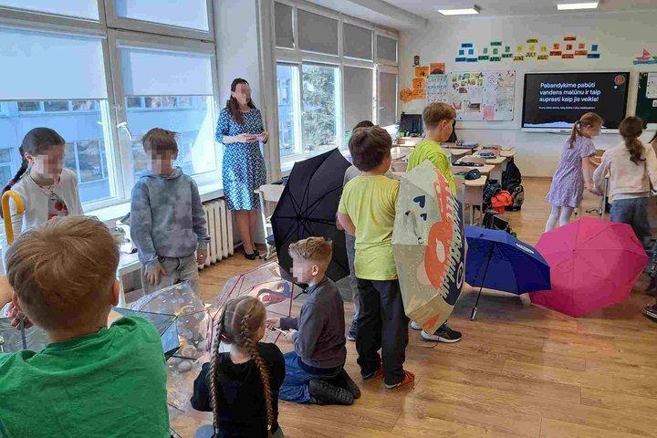 Nuotraukoje – pradinukai klasėje, dalyvaujantys pamokoje apie vandens malūną. Vaikai su skėčiais vaidina malūno mechanizmus. Prie lango stovi mokytoja, o lentoje parašyta: „Pabandykime pabūti vandens malūnu ir taip suprasti kaip jis veikia!“ Aplinka kūryb