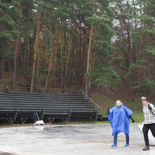 Du vyrai eina per lauko estrados aikštelę, vienas jų tempia didelį baltą šiukšlių maišą, o kitas vilki ryškų mėlyną lietpaltį ir rodo pergalės ženklą. Fone matyti mediniai žiūrovų suoliukai ir miškingas šlaitas su aukštais pušynais. Aplinka drėgna, dangus