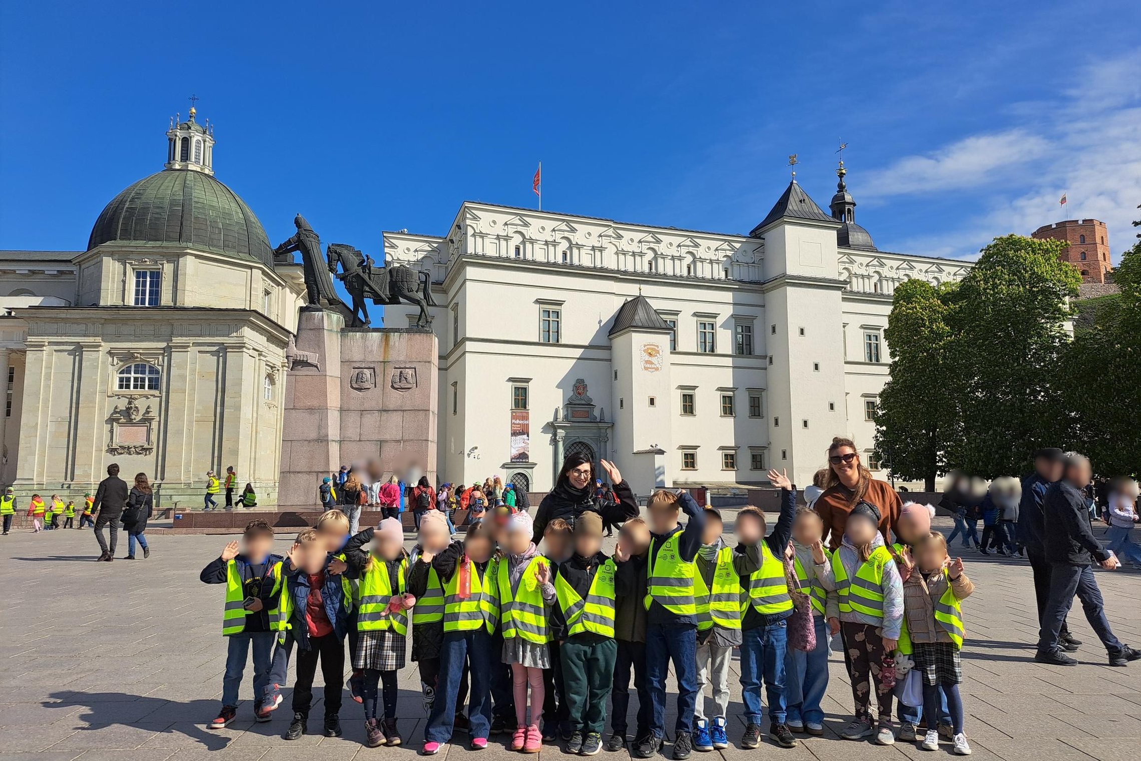 Nuostabi nuotrauka! Vaikai, pasipuošę ryškiomis liemenėmis, stovi priešais Lietuvos Didžiosios Kunigaikštystės valdovų rūmus Vilniuje. Fone matyti didingas paminklas kunigaikščiui Gediminui su žirgu, o tolumoje – Gedimino pilies bokštas. Saulėtas oras ir