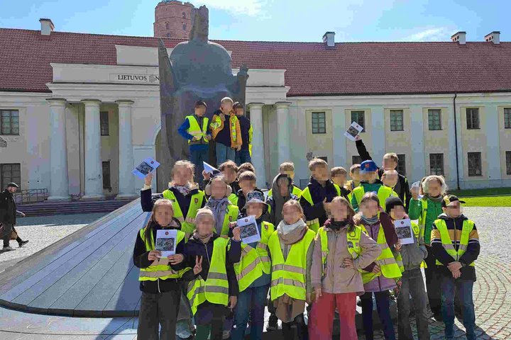 Ketvirtokų grupė po edukacijos „Žemutinės pilies paslaptys“ Vilniaus pilių teritorijoje, pozuoja prie karaliaus Mindaugo paminklo. Vaikai vilki ryškias liemenes ir rankose laiko edukacijos užduotis.