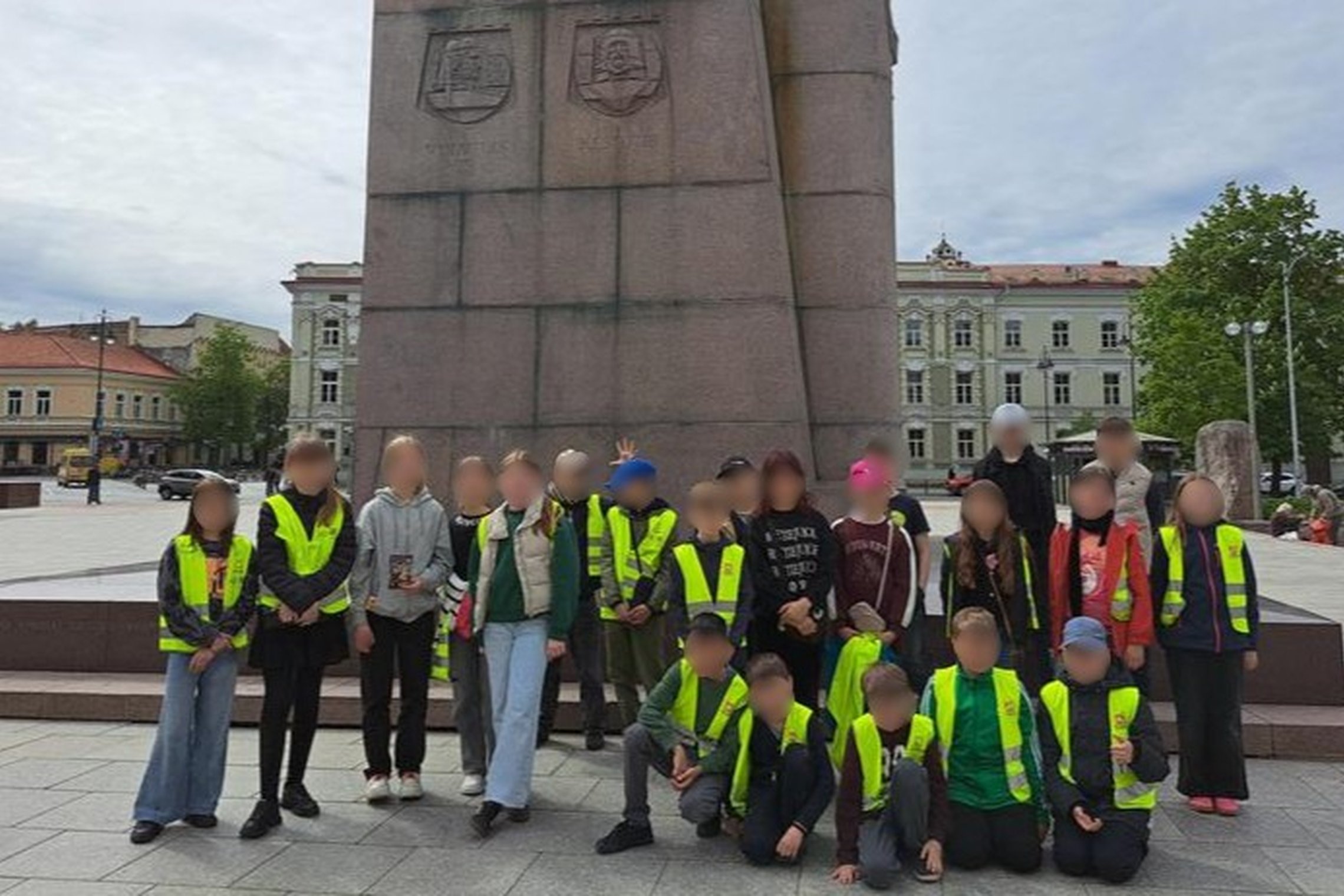 Nuotraukoje matyti vaikų grupė, apsirengusi ryškiai geltonomis liemenėmis – tikėtina, tai mokiniai išvykos metu. Jie pozuoja prie paminklo su herbais – tai yra paminklas didžiajam kunigaikščiui Gediminui Vilniuje, Katedros aikštėje. Fone matyti istoriniai