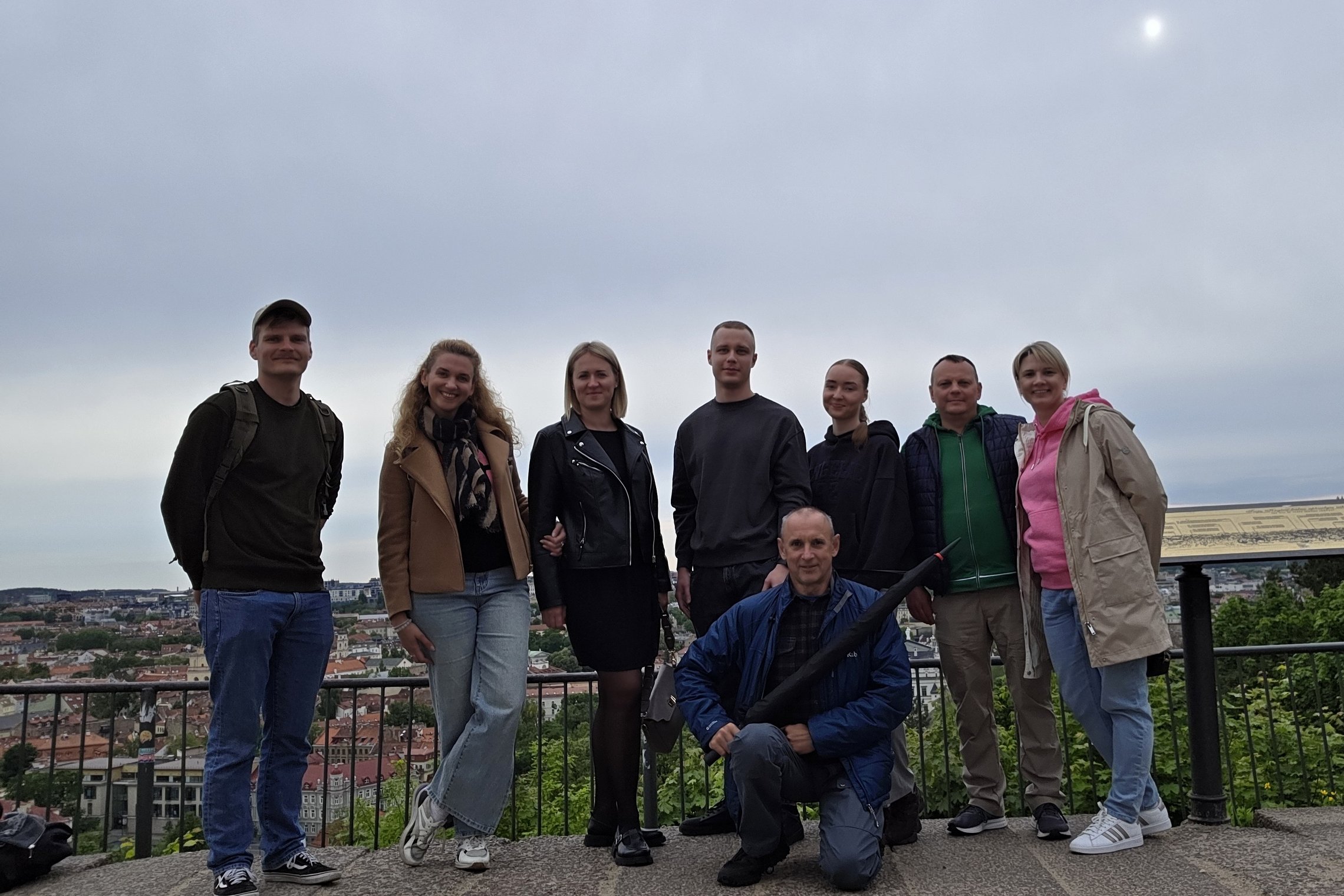 Grupė Pasieniečių mokyklos atstovų stovi ant Trijų Kryžių kalno apžvalgos aikštelės Vilniuje, už jų atsiveria miesto panorama; priekyje – ekskursijos vadovas su skėčiu rankoje.