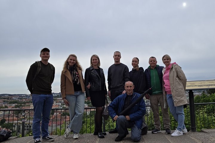 Grupė Pasieniečių mokyklos atstovų stovi ant Trijų Kryžių kalno apžvalgos aikštelės Vilniuje, už jų atsiveria miesto panorama; priekyje – ekskursijos vadovas su skėčiu rankoje.