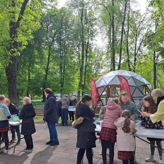 Vaikai susibūrę prie kelių stalų dalyvauja edukacinėje veikloje lauke, apsupti vešlios žalumos. Fone matyti stiklinis kupolas su dviem raudonomis vėliavomis. Vaikus stebi ir padeda kelios mokytojos. Aplinka – pavasariškai žaliuojantis parkas su suoliukais
