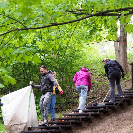 Grupė žmonių kopia mediniais laiptais miške, nešdami didelį baltą maišą ir valydami pakraščius. Žali medžių lapai virš jų sudaro natūralų stogą.