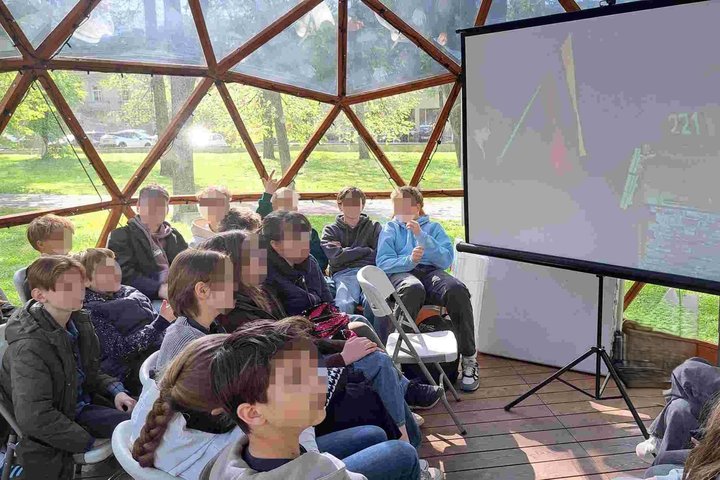 Mokiniai susidomėję stebi vaizdo medžiagą apie „Roko maršą per Lietuvą“ edukacijoje kupole. Edukacinė erdvė po atviru dangumi kuria jaukią ir įtraukiančią mokymosi atmosferą.