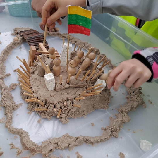 Nuotraukoje matyti vaikų sukurta piliakalnio maketo instaliacija. Ji pagaminta iš smėlio, medinių pagaliukų, putplasčio detalių bei kitų kūrybinių priemonių. Maketas vaizduoja įtvirtintą piliakalnį su vartais, tiltu, sargybiniais bei Lietuvos vėliava cent