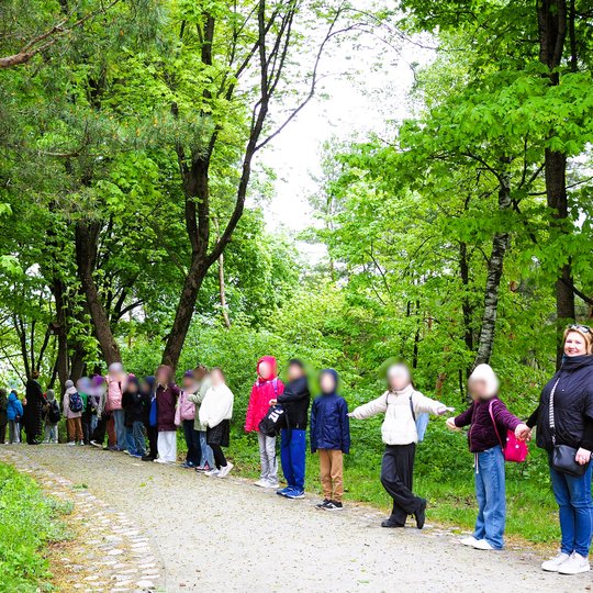 Vaikai ir mokytoja stovi ilga gyva grandine miško takelyje. Jie susikibę rankomis tarp medžių žalumos, šypsosi ir žvelgia į kamerą.