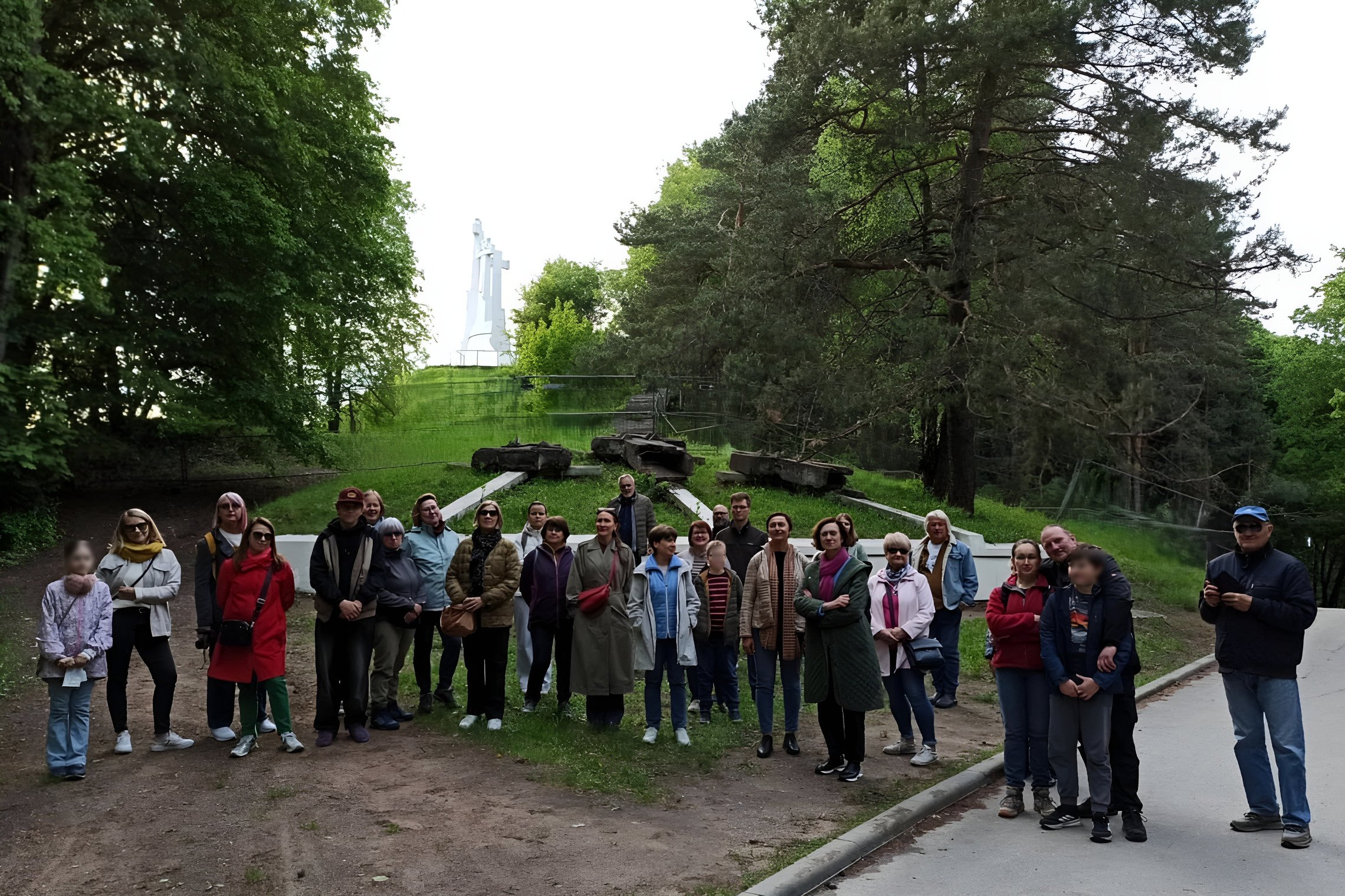 Grupė žmonių stovi Kalnų parke, Vilniuje. Fone matomas Trijų kryžių paminklas ant kalno, aplink žali medžiai. Priekyje – susprogdintų senųjų Trijų Kryžių betoninės konstrukcijos, ekskursijos dalyviai pozuoja bendrai nuotraukai. Atmosfera rami, vakarinė šv
