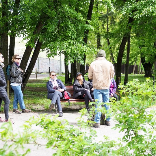 Grupė žmonių parke klausosi vyro, stovinčio priešais juos ir kažką pasakojančio. Kai kurie sėdi ant suoliuko, kiti stovi, o aplink žaliuoja medžiai ir krūmai.